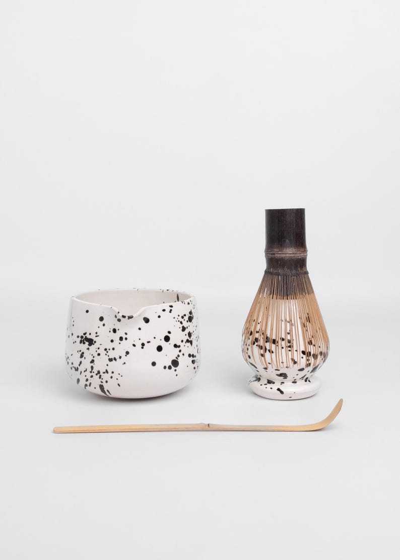 Puede incluir: Un cuenco de matcha de cer&aacute;mica blanca con un dise&ntilde;o de pintura salpicada negra, un batidor de matcha de bamb&uacute; con una base de cer&aacute;mica blanca con un dise&ntilde;o de pintura salpicada negra, y un batidor de matcha de bamb&uacute;.