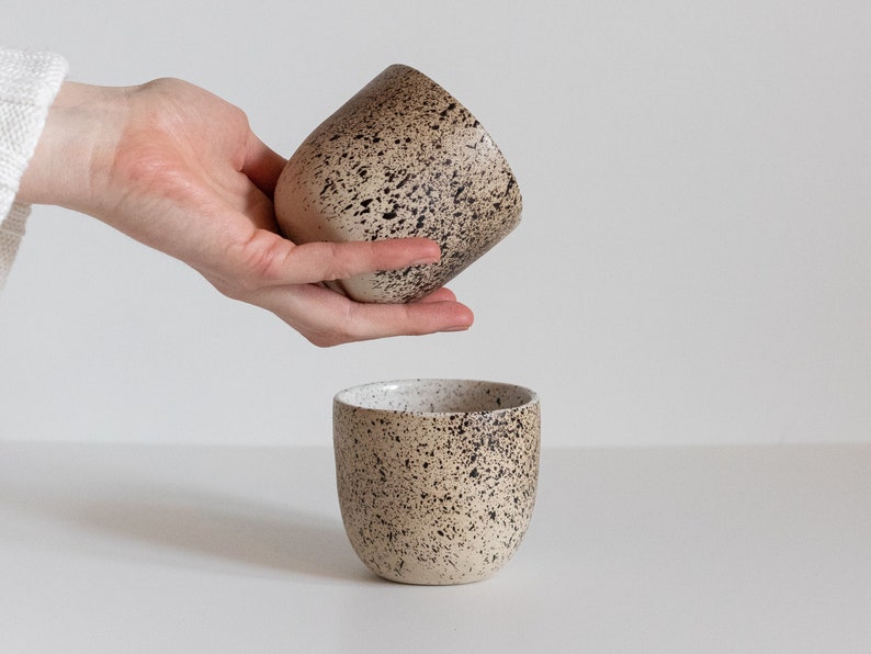 May include: Two speckled ceramic mugs, one in a hand and the other on a white surface. The mugs are a light beige color with dark brown speckles.