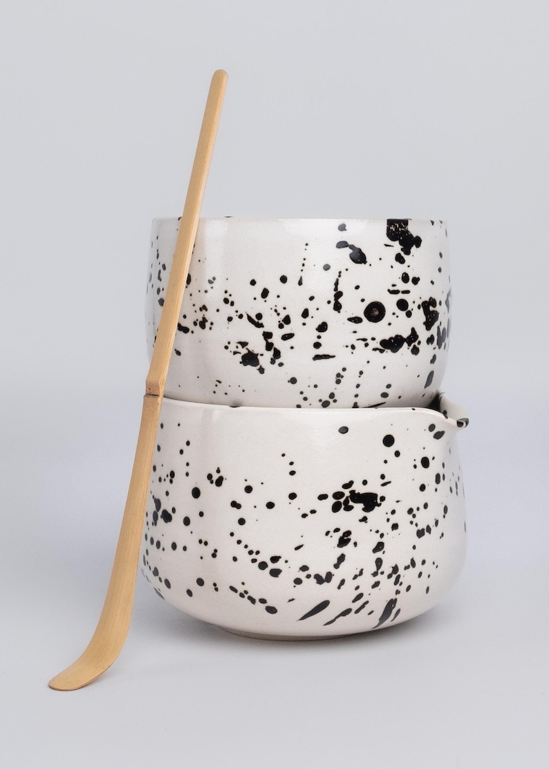Puede incluir: Dos cuencos de cer&aacute;mica blanca con un dise&ntilde;o de pintura negra salpicada, apilados uno encima del otro. Un batidor de matcha de madera se coloca al lado de los cuencos.