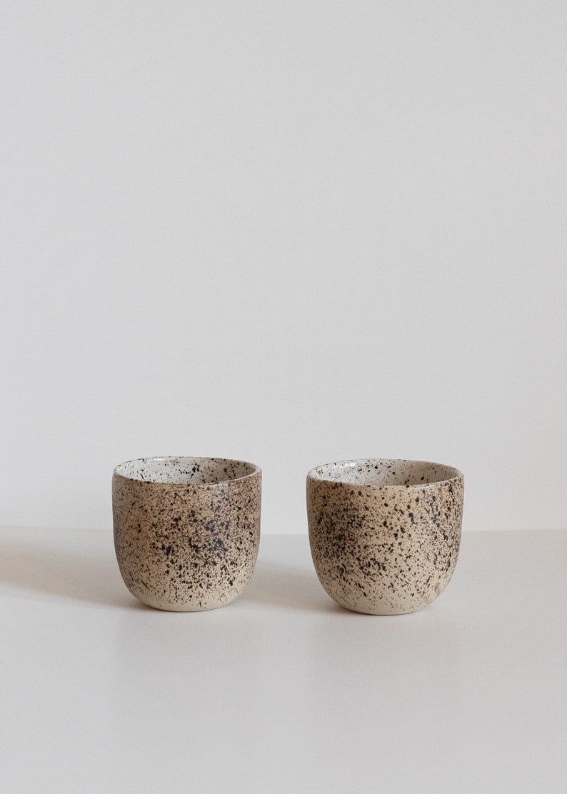 May include: Two speckled ceramic cups with a white and brown glaze. The cups are sitting on a white surface.