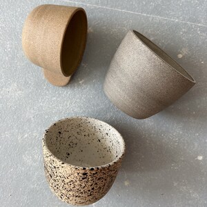 May include: Three ceramic mugs in different colors and patterns. The first mug is a light brown with a speckled texture. The second mug is a light gray with a speckled texture. The third mug is white with black speckles.