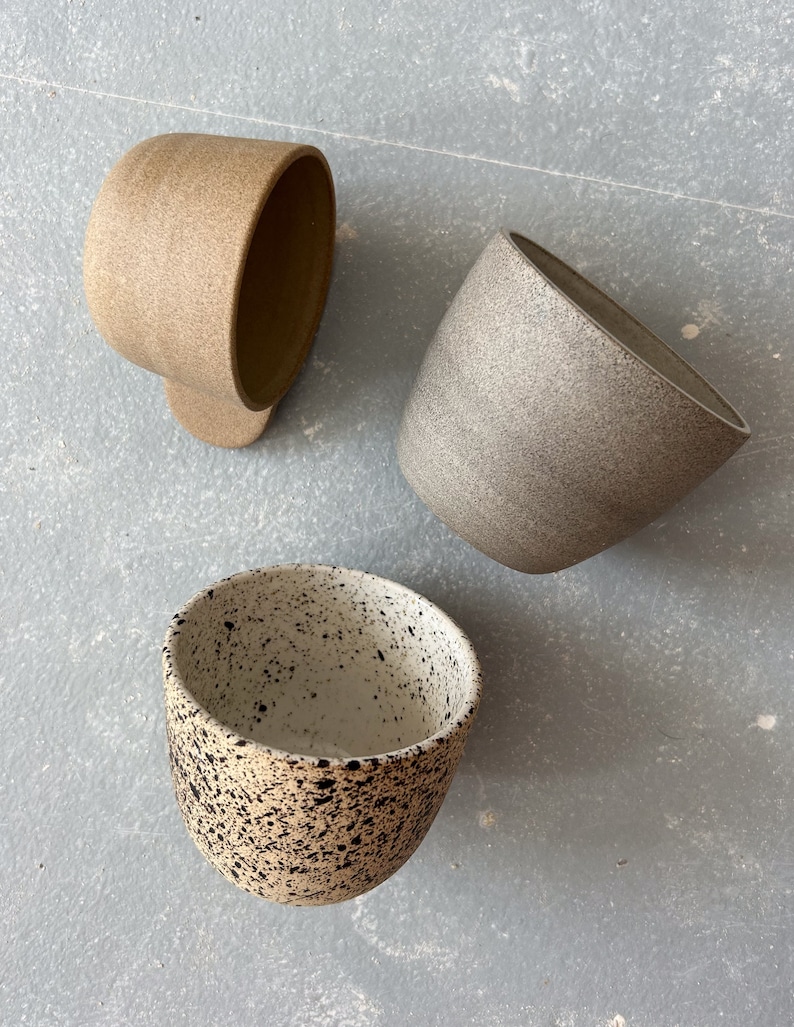 May include: Three ceramic mugs in different colors and patterns. The first mug is a light brown with a speckled texture. The second mug is a light gray with a speckled texture. The third mug is white with black speckles.