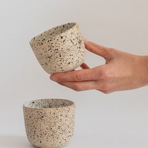 May include: Two speckled beige ceramic mugs with black speckles. The mug on top is being held by a hand.