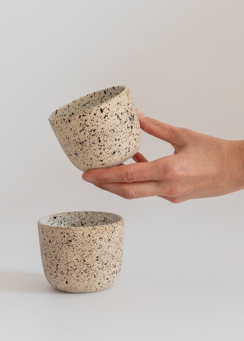 May include: Two speckled beige ceramic mugs with black speckles. The mug on top is being held by a hand.