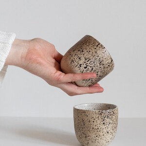 May include: Two speckled ceramic mugs, one in a hand and the other on a white surface. The mugs are a light beige color with dark brown speckles.