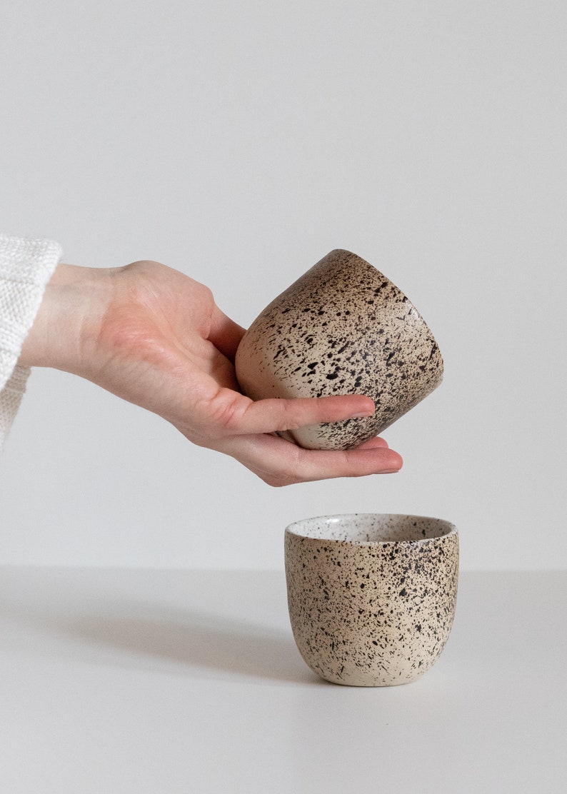 May include: Two speckled ceramic mugs, one in a hand and the other on a white surface. The mugs are a light beige color with dark brown speckles.