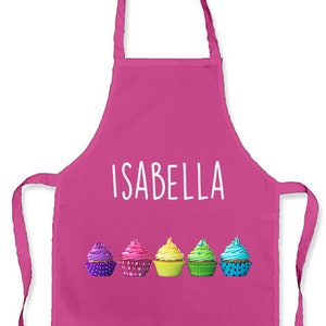 May include: Pink apron with the name "ISABELLA" printed in white. Five colourful cupcakes are lined up across the bottom of the apron.