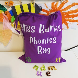 May include: Purple fabric bag with white text that reads "Miss Burke's Phonics Bag". The bag is sitting on a white surface with colourful letter tiles scattered around it.