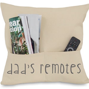 May include: A beige throw pillow with a pocket on the front. The pocket has the words "dad's remotes" embroidered on it. A remote control and a stack of magazines are in the pocket.
