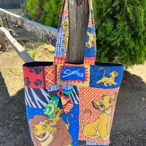 May include: A fabric tote bag featuring a patchwork design with various patterns and images of Simba from The Lion King. The bag includes a blue rectangular patch with the text Simba, a small decorative bow, and two shoulder straps.