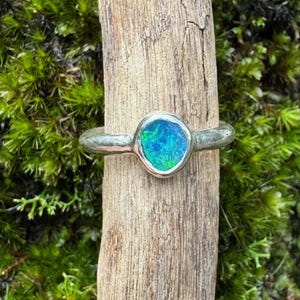 May include: A silver ring with a round, iridescent blue and green stone. The ring is set on a piece of wood against a backdrop of green moss.