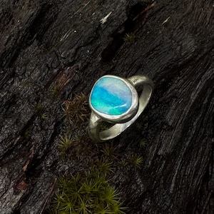 Puede incluir: Un anillo de plata con una piedra de ópalo ovalada. El ópalo muestra una vibrante mezcla de tonos azules y verdes. El anillo se encuentra sobre un fondo oscuro y texturizado, resaltando los colores de la piedra preciosa.