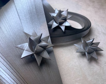 Puede incluir: Primer plano de tres estrellas de origami plateadas, hechas con tiras finas de papel. Las estrellas están dispuestas sobre una superficie blanca, con una estrella parcialmente oculta por un rollo de papel plateado.