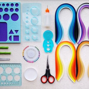 May include: A set of paper quilling tools and supplies, including a variety of coloured paper strips, a quilling tool, a ruler, a circle template, a pincushion, a bottle of glue, and a pair of scissors.