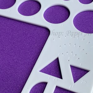 May include: White plastic circle and triangle template with various sizes marked in inches. The template is on a purple textured surface. The text "Shop: Paperyuan" is visible in the upper right corner.