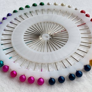 May include: A white plastic pincushion with a circular base and a central hub with radiating spokes. The pincushion is filled with colourful pearl-like beads in a rainbow pattern. The text "Shop: Paper Art Junky" is visible on the base.