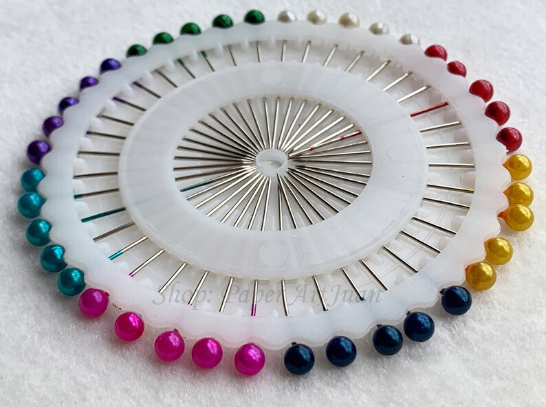 May include: A white plastic pincushion with a circular base and a central hub with radiating spokes. The pincushion is filled with colourful pearl-like beads in a rainbow pattern. The text "Shop: Paper Art Junky" is visible on the base.