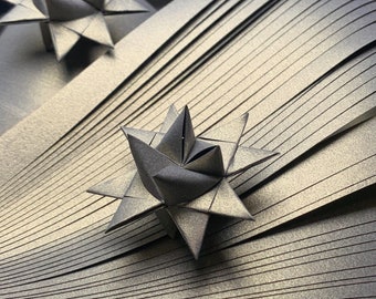 Puede incluir: Dos estrellas de papel negro, dobladas en un estilo origami tradicional, se colocan sobre un fondo de tiras de papel dorado. Las estrellas se superponen, creando un efecto tridimensional.