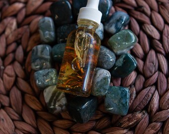 May include: A clear glass bottle with a white dropper filled with amber liquid, resting on a bed of green and gray stones. The stones are arranged on a woven brown surface, creating a natural and earthy aesthetic.