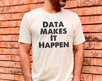 May include: A person wearing a light beige t-shirt with the text "DATA MAKES IT HAPPEN" printed in black. The person is standing in front of a brick wall.