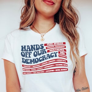 May include: White t-shirt with a blue and red graphic that says "Hands Off Our Democracy" with a list of items under it, including "Free Speech", "Federal Programs", "Healthcare", "DEI", "Students", "Medicaid", "Medicare", "Federal Workers", "Scientists", "National Parks", "Courts", "Immigrants", "Personal Data", "Our Rights", "Fair Elections", "Education", and "Social Security".