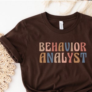 May include: A brown t-shirt with the text "BEHAVIOR ANALYST" in colorful retro lettering.