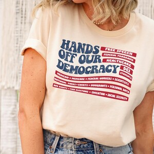 May include: A beige t-shirt with a red, white, and blue American flag design. The text "Hands Off Our Democracy" is printed in blue on the flag. The text "Free Speech, Federal Programs, Healthcare, DEI, Students, Medicaid, Medicare, Federal Workers, Scientists, National Parks, Courts, Immigrants, Personal Data, Our Rights, Fair Elections, Education, Social Security" is printed in black below the flag.