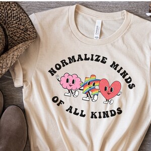 May include: A beige t-shirt with the text "Normalize Minds of All Kinds" in black. The text is surrounded by a cartoon brain, jigsaw piece, and heart.