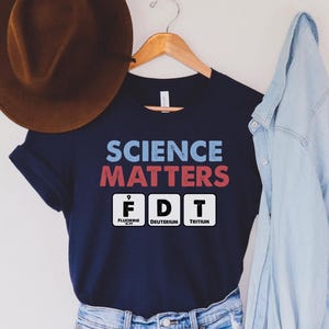 May include: Navy blue t-shirt with the text "SCIENCE MATTERS" in blue and red, and the chemical symbols F, D, and T. A brown hat and a light blue denim jacket are in the background.