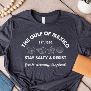 May include: Heather navy t-shirt with white text that reads "THE GULF OF MEXICO EST. 1550 STAY SALTY & RESIST fresh dreamy tropical" and seashell graphics. The shirt is knotted at the bottom and has a round neckline.