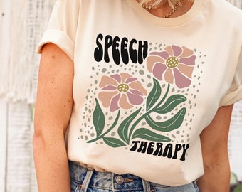 May include: A light beige t-shirt with a graphic design of two pink flowers and green leaves. The text "SPEECH THERAPY" is printed in black letters on the shirt.