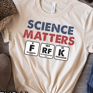 May include: A beige t-shirt with the words "SCIENCE MATTERS" in blue and red. Below the text are three periodic table element boxes: Fluorine, Rutherfordium, and Potassium. The shirt is tied at the waist.