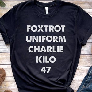 May include: Black t-shirt with white text: "Foxtrot Uniform Charlie Kilo 47". The word "BLACK" is printed at the bottom. The shirt is slightly wrinkled and has rolled-up sleeves. Part of a brown hat and blue jeans are visible.