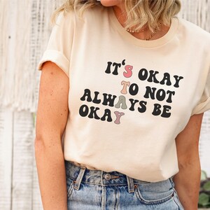 May include: A light beige t-shirt with a black graphic that reads "It's okay to not always be okay". The graphic has colorful letters in shades of pink, green, and brown.