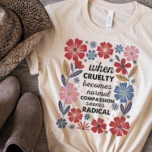May include: A natural-colored t-shirt with the text "when cruelty becomes normal compassion seems radical" surrounded by colorful flowers. The flowers are red, pink, blue, and green. The shirt is knotted at the bottom.