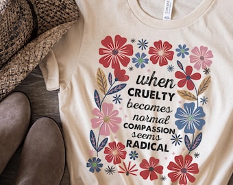 May include: A natural-colored t-shirt with the text "when cruelty becomes normal compassion seems radical" surrounded by colorful flowers. The flowers are red, pink, blue, and green. The shirt is knotted at the bottom.