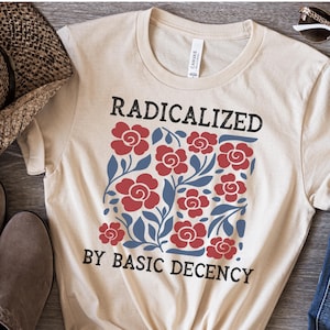 May include: A natural-colored t-shirt with the text "RADICALIZED BY BASIC DECENCY" in black. The shirt features a floral design with red roses and blue leaves. The shirt is knotted at the bottom.