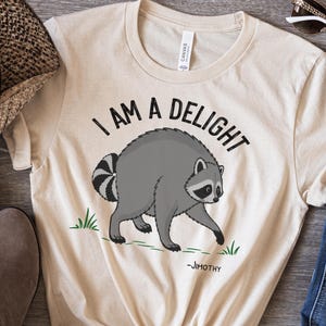 May include: A natural-colored t-shirt featuring a graphic of a gray raccoon with a striped tail. The text above the raccoon reads "I AM A DELIGHT" in black capital letters, and the name "-JIMOTHY" is printed in smaller black text below the raccoon.