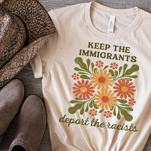 May include: A cream-coloured t-shirt with the slogan "KEEP THE IMMIGRANTS deport the racists" in green. The shirt has a floral design with orange and yellow flowers and green leaves. The word "NATURAL" is at the bottom left.