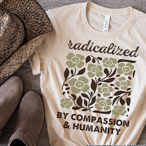 May include: A cream-coloured t-shirt with the words "radicalized" and "BY COMPASSION & HUMANITY" printed on it. The shirt features a floral design in shades of green and brown. The word "NATURAL" is printed on the bottom left.