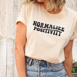May include: A cream coloured t-shirt with the text "Normalize Positivity" in black, retro-style lettering.