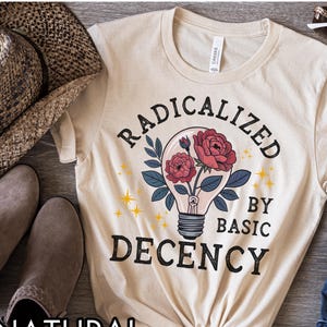 May include: A natural-colored t-shirt with the text "RADICALIZED BY BASIC DECENCY" in black. The design includes a lightbulb with red roses and green leaves. The word "NATURAL" is printed at the bottom.
