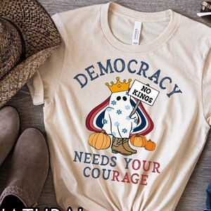 May include: A natural-colored t-shirt featuring a cartoon ghost wearing a crown and cowboy boots, holding a sign that reads "NO KINGS." The shirt also displays the words "DEMOCRACY NEEDS YOUR COURAGE" in blue and red.