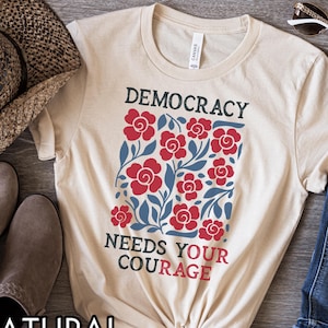 May include: A natural-colored t-shirt with the words "DEMOCRACY NEEDS YOUR COURAGE" in black and red. The shirt features a floral design with red flowers and blue leaves. The shirt is knotted at the bottom.