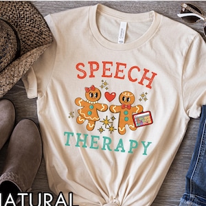 Peut inclure: Un t-shirt beige avec les mots "SPEECH THERAPY" en rouge et turquoise. Le t-shirt présente des bonhommes en pain d'épices de dessin animé se tenant la main, avec un cœur et des étoiles. Le mot "NATURAL" est imprimé en bas.