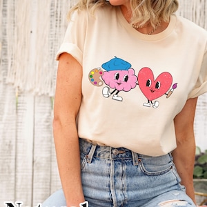 Peut inclure: Un t-shirt crème avec un motif représentant un cerveau rose portant un béret bleu et tenant une palette de peinture, et un cœur rouge tenant un pinceau. Le texte "Natural" est imprimé en bas de l'image.