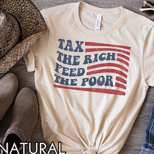 May include: A natural-colored t-shirt with the text "TAX THE RICH FEED THE POOR" in blue and red, designed to resemble a flag. The shirt is made of a soft material and is suitable for casual wear.