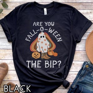 May include: Black t-shirt with a Halloween-themed graphic. The design features a ghost wearing cowboy boots, surrounded by pumpkins and a rainbow arch. Text reads "ARE YOU FALL-O-WEEN THE BIP?"