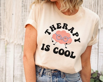 May include: A natural-colored t-shirt with the text "THERAPY IS COOL" in black, arched above a cartoon brain wearing glasses. The brain has a smiling face and arms and legs. The word "Natural" is at the bottom.