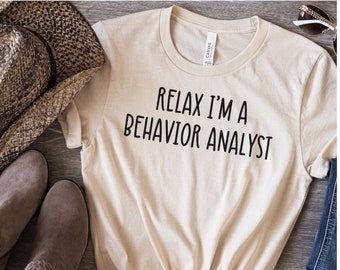 May include: A beige t-shirt with the text "Relax I'm a Behavior Analyst" printed in black. The shirt is laid out on a wooden surface with a pair of brown boots and a straw hat.
